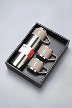 A stainless steel vacuum flask bottle with two matching ceramic cups presented in a black gift box. The bottle has a silver metallic body with a beige handle and collar, and a white and red paper label.