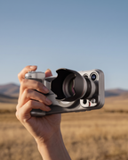 A hand holding the grip and lens outdoors against a blurred field and blue sky