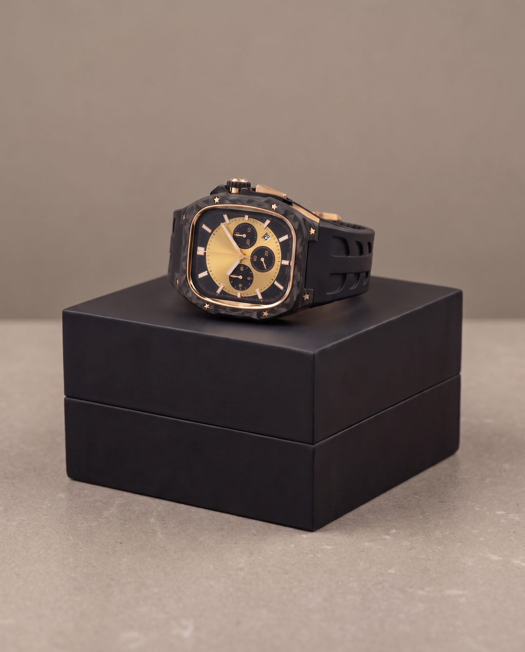 A luxury watch with a black carbon fiber case, gold-toned dial, and black strap, displayed on top of a black presentation box against a neutral beige background.