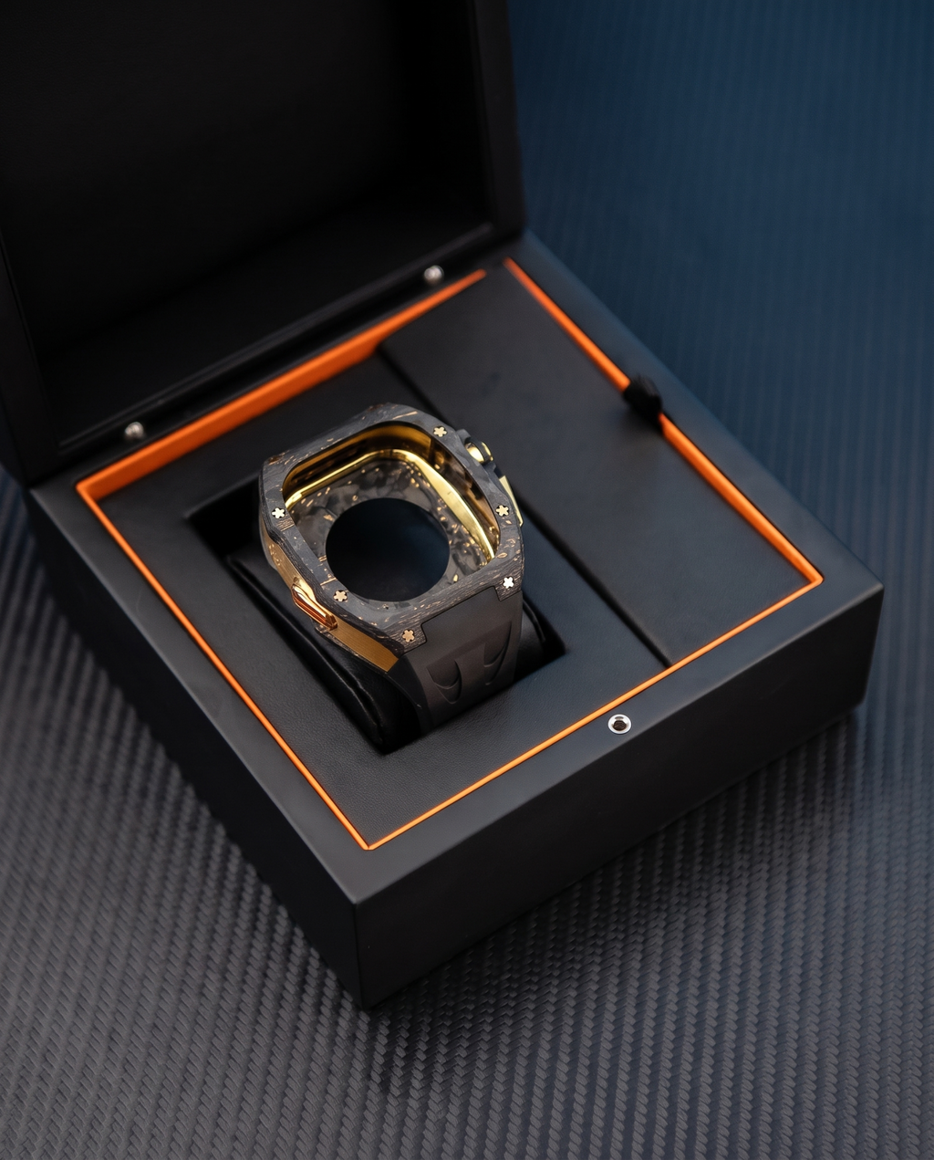 A luxury watch with a gold and black bezel and a dark gray carbon fiber strap, presented inside an open black watch box with orange lining on a textured dark gray surface.