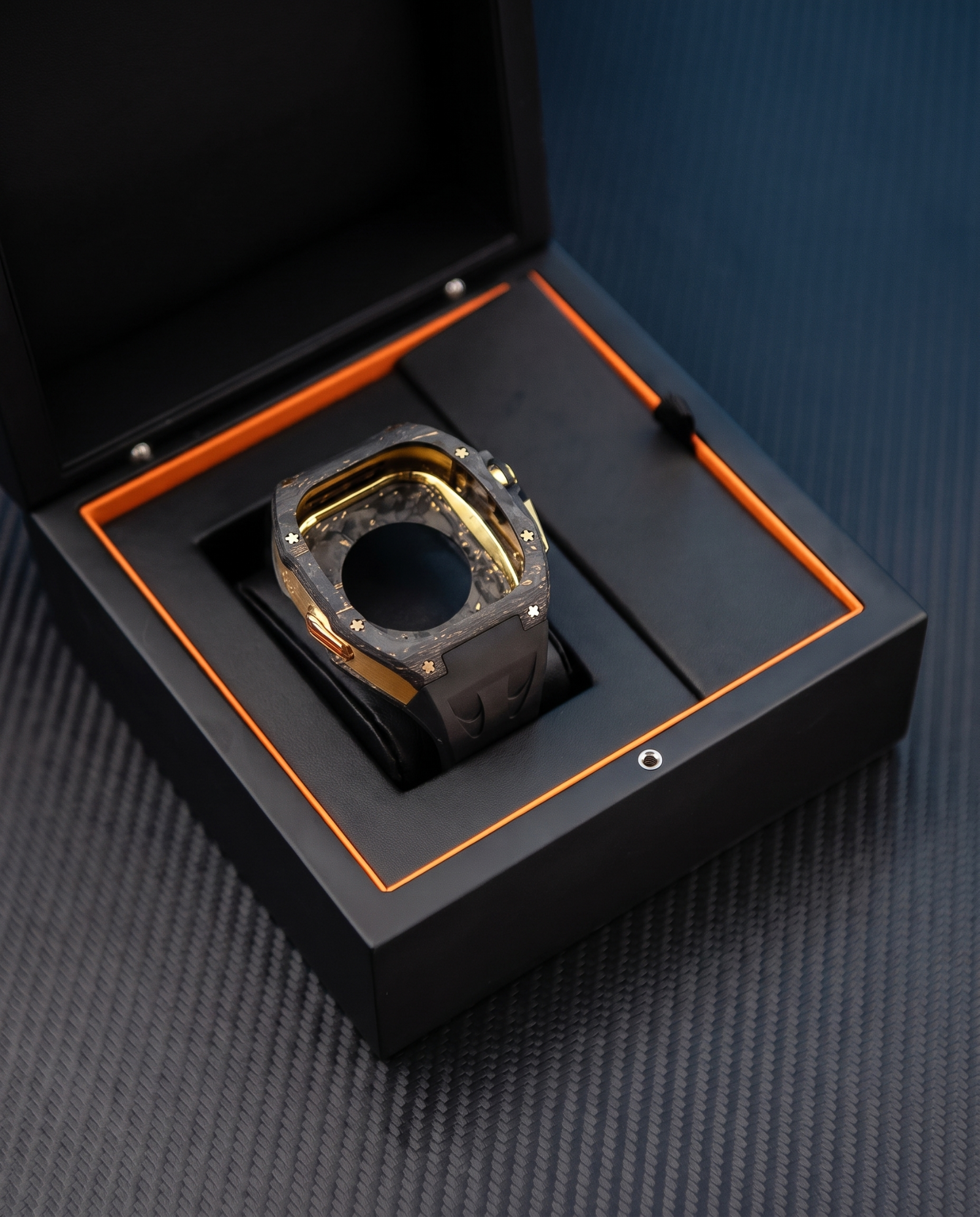 A luxury watch with a gold and black bezel and a dark gray carbon fiber strap, presented inside an open black watch box with orange lining on a textured dark gray surface.
