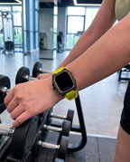 Woman in a gym wearing the case kit on a chartreuse strap, forearm resting on a dumbbell rack