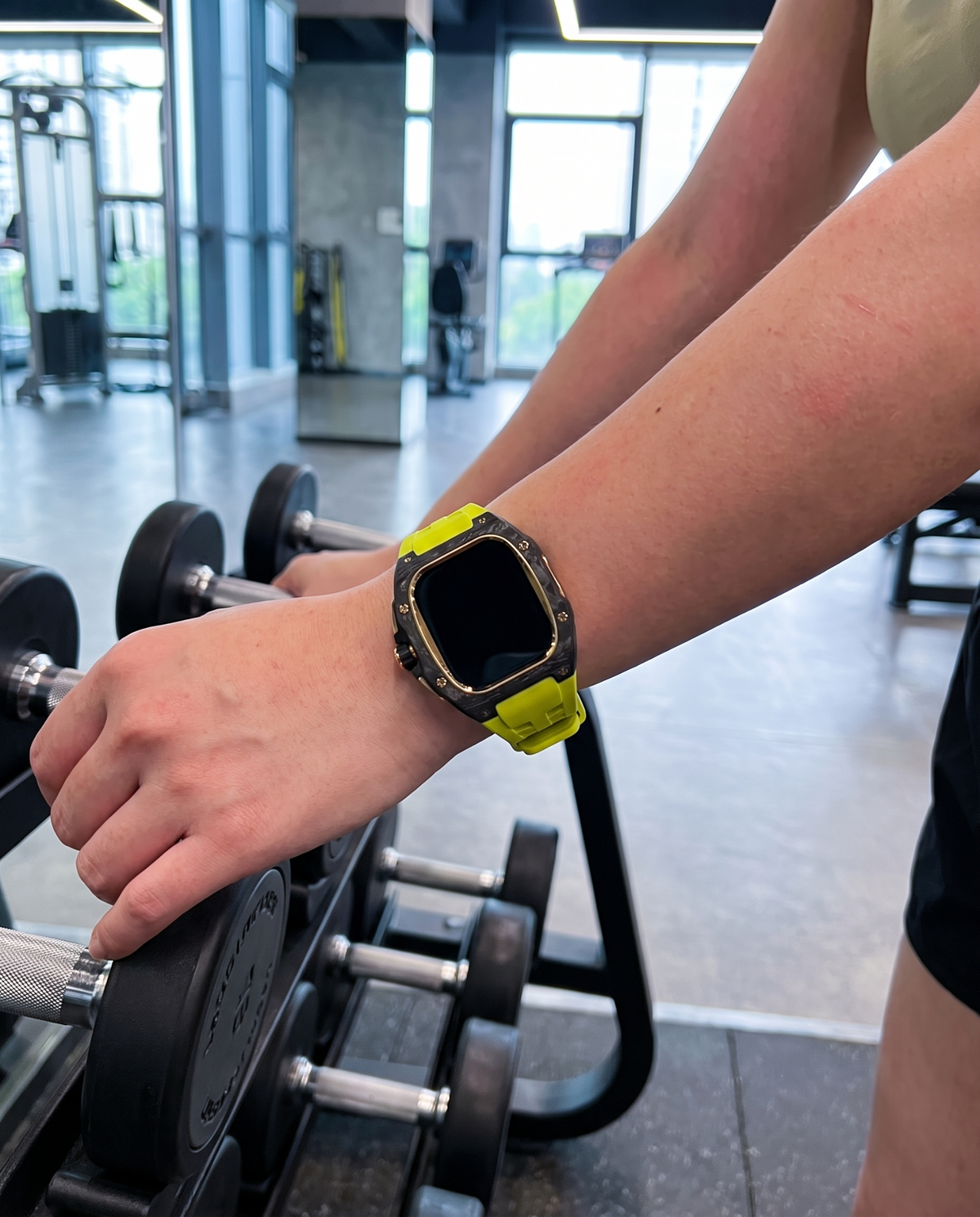 Woman in a gym wearing the case kit on a chartreuse strap, forearm resting on a dumbbell rack