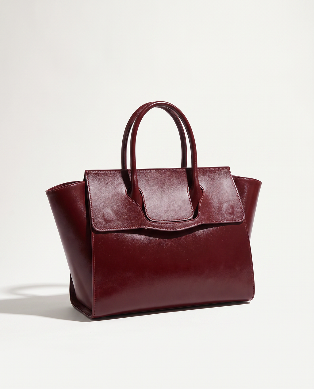 A burgundy leather handbag with double top handles and a front flap, photographed on a clean white studio background.