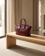 Wine red full grain leather laptop tote resting on a pale oak bench in a calm sunlit interior