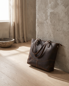 Dark brown crazy horse leather tote resting against a stone wall in a calm sunlit interior