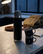 The flask and nested cups on a stone worktop before dawn, beside a half-packed daypack