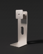 Silver anodised aluminium monitor stand with a weighted base and an oval cable slot cut through the column, shown against a graphite background.