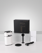 A countertop coffee grinder set displayed on a light gray studio surface, including a white cylindrical grinder body, a black lid, a black stand, a black power cord, a black instruction booklet, and a black product box with a line drawing of the grinder.