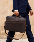 A man in a navy blue suit carrying a dark brown leather briefcase with a top handle and adjustable shoulder strap, set against a textured beige wall.
