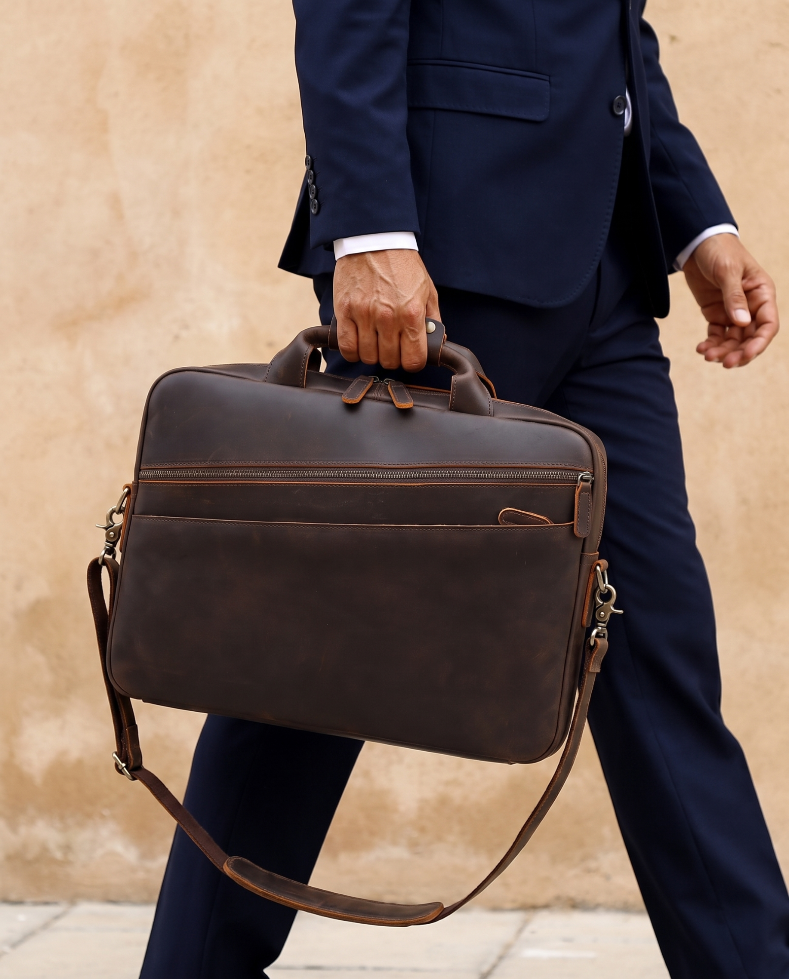 A man in a navy blue suit carrying a dark brown leather briefcase with a top handle and adjustable shoulder strap, set against a textured beige wall.