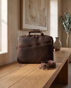 A dark brown leather briefcase with a matching adjustable shoulder strap, styled on a rustic wooden table in a warm, sunlit room.