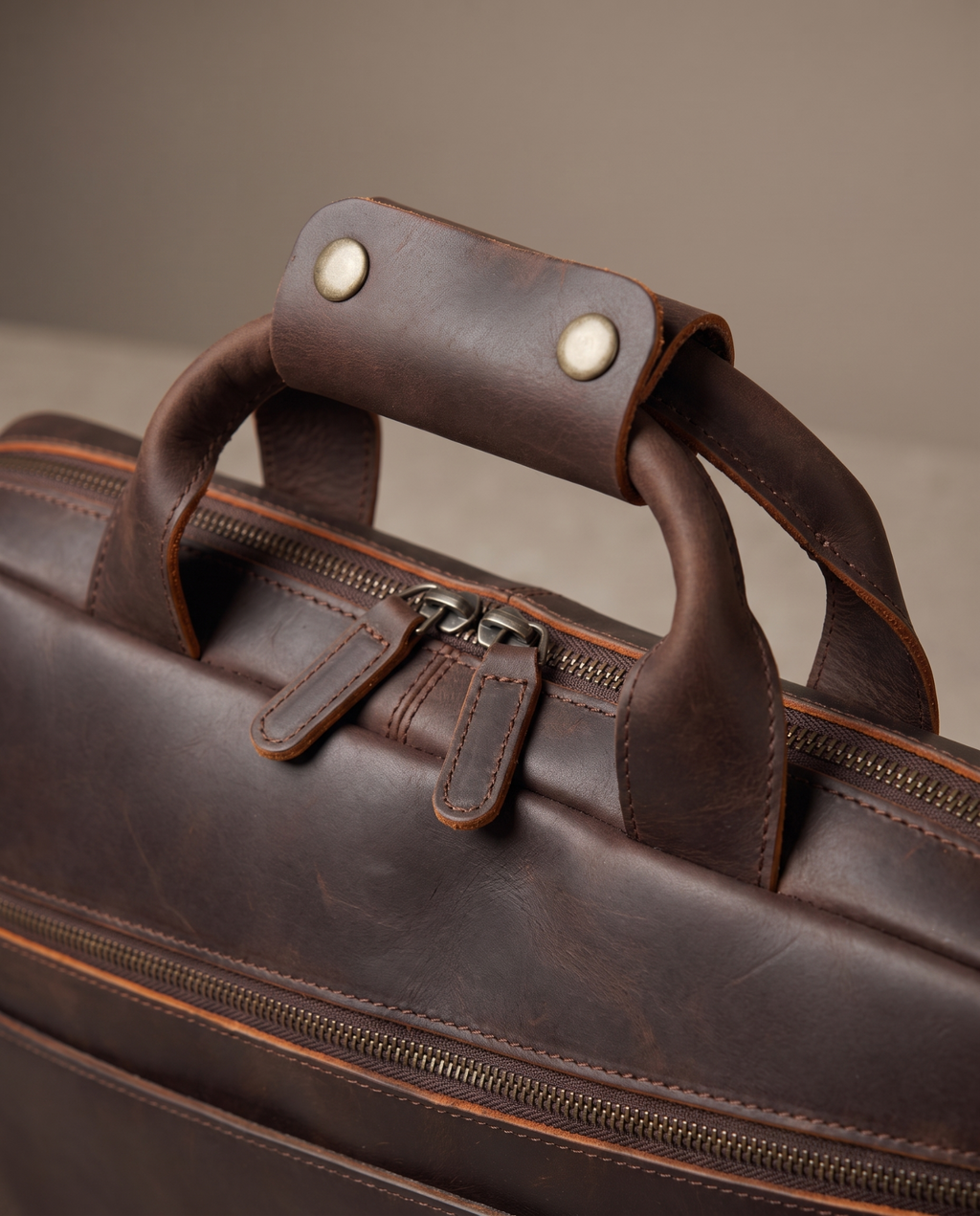 Close-up of a dark brown leather briefcase or duffel bag with a top handle, zipper closure, and metal snap details, photographed against a neutral beige background.