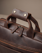 Close-up of a dark brown leather briefcase or duffel bag with a top handle, zipper closure, and metal snap details, photographed against a neutral beige background.