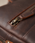 Close-up of a dark brown leather bag showing a zipper with an antique brass pull and detailed stitching.