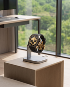 A luxury watch with a gold skull dial and black strap, displayed on a modern silver charging stand on a light wood pedestal in a bright office setting.